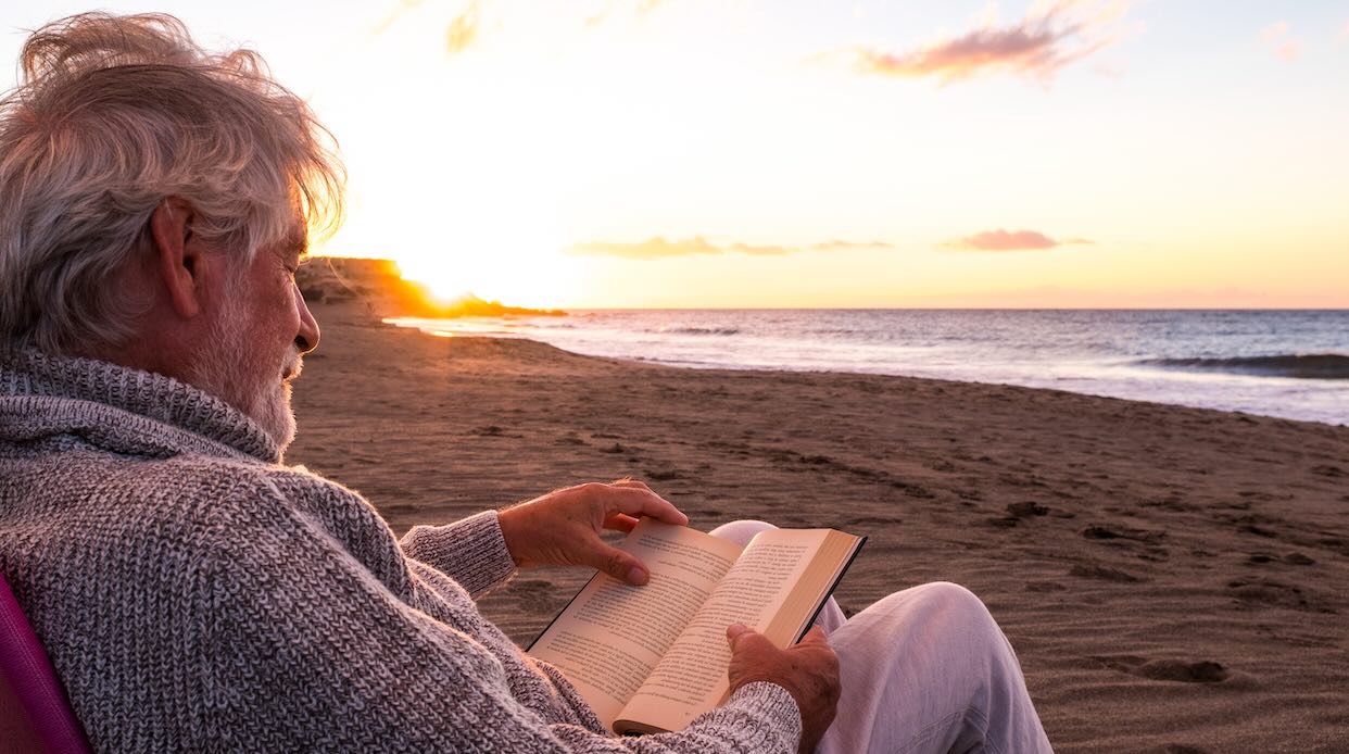A mature man sits reading a book along a quiet, deserted shoreline, reflecting on how familiar words can take on new meaning with time.