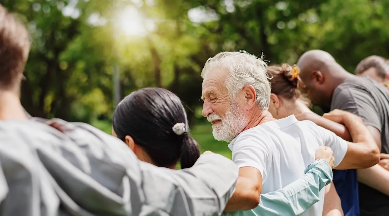 A diverse group of adults standing together outdoors with their arms around one another, including an older man at the center.